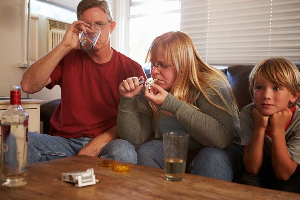 dad drinking while mom lights a pipe and child sits as drug-addicted parents affect him