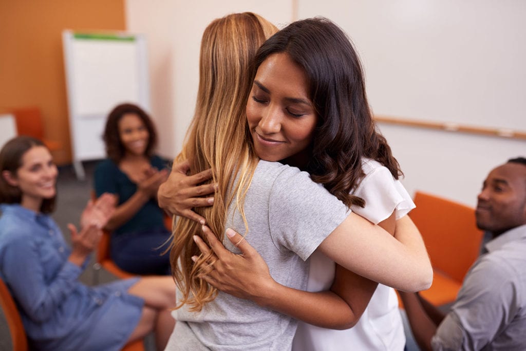 two women hug after one explains to a group how a 12 step program helps recovery